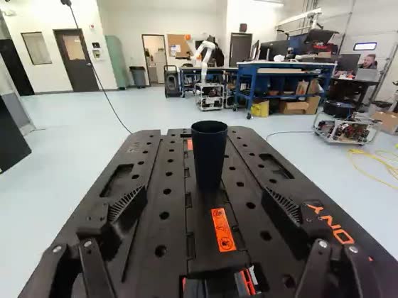 Wrist camera: the gripper descends toward a dark cup on a slotted black workbench, the laboratory behind it.