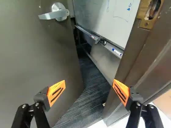 Wrist camera: the gripper is level with the lever handle of a heavy grey door standing ajar, carpeted floor and a whiteboard visible through the gap.