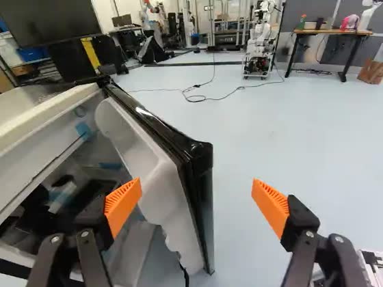 Wrist camera: the gripper grips the edge of an opened white refrigerator door; an open laboratory with benches and a second robot lies beyond it.